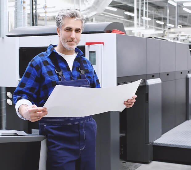 Zadowolony mężczyzna w stroju roboczym stojący na hali produkcyjnej, trzymający w rękach duży arkusz papieru. Za mężczyzna stoi duża maszyna produkcyjna